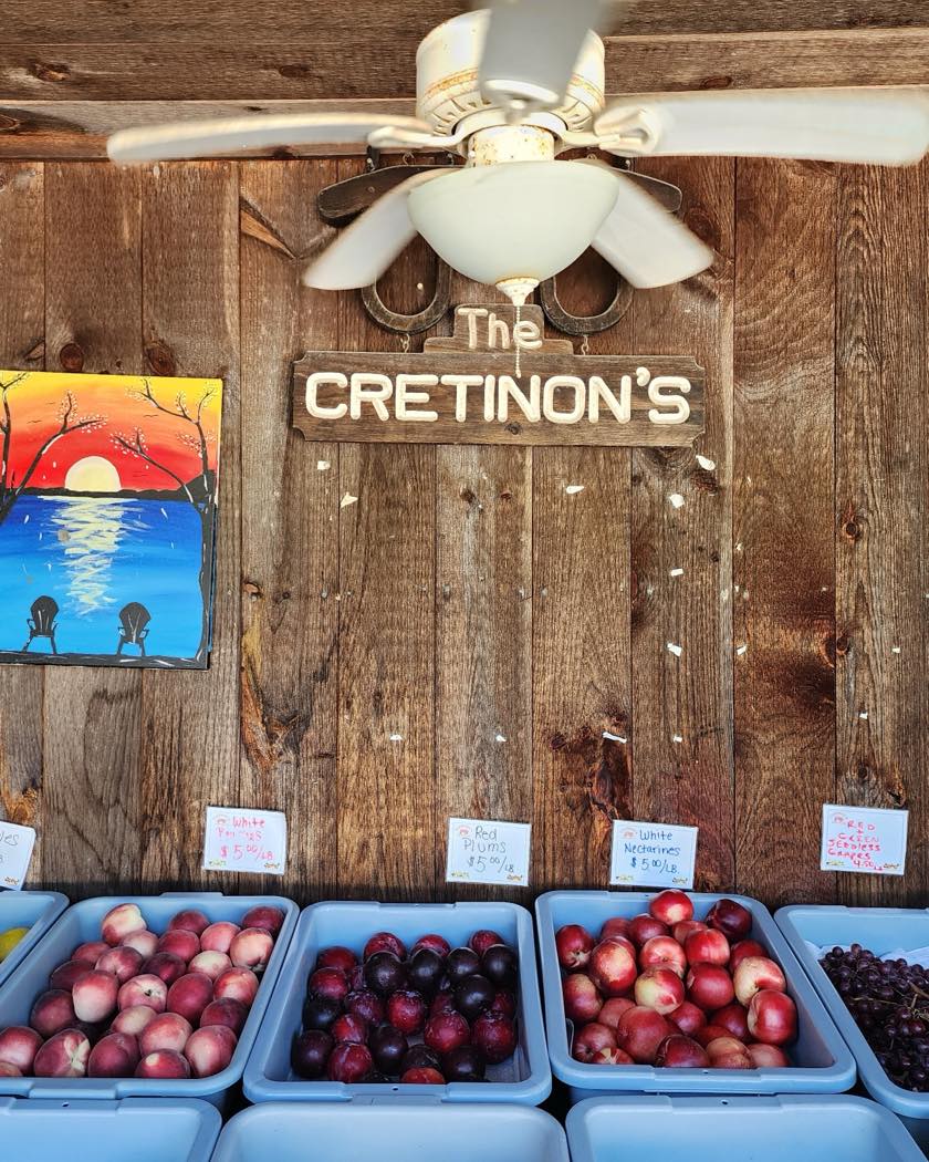 The hand-painted "Cretinon's" sign and ceiling fan above baskets of fresh peaches, plums, and grapes inside the farmstand